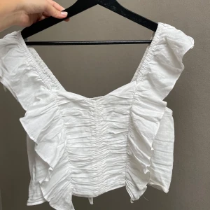 Vit linnetopp med volanger - Säljer en superfin vit linnetopp i linneblandning. Toppen har volanger längs axlarna och en smockad design som ger en snygg passform. Perfekt för sommardagar eller en festlig kväll!