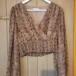 Ormmönstrad omlottopp från NA-KD - Säljer en supercool ormmönstrad omlottopp från NA-KD. Den har långa ärmar och en croppad passform som ger en trendig look. Perfekt för en utekväll eller när du vill sticka ut lite extra. Toppen är i ett mjukt och bekvämt material.