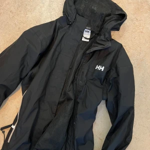 Svart regnjacka från Helly Hansen - Säljer en svart regnjacka från Helly Hansen. Den har en praktisk huva och dragkedja framtill. Perfekt för regniga dagar och håller dig torr och bekväm. Passar både till vardags och utomhusaktiviteter. Som ny.
