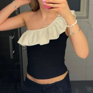 Svart-vit oneshoulder topp från H&M - Snygg svart oneshoulder topp med vit volangdetalj. Toppen är ärmlös och har en elegant asymmetrisk design. Bara använd 2 gånger. 💕