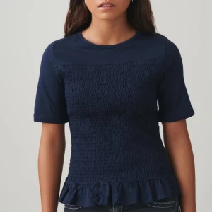 Mörkblå topp med volangdetalj - Snygg mörkblå topp med korta ärmar och volangdetalj vid nederkanten. Toppen har en smockad textur på framsidan som ger en unik look. Perfekt för en stilren och chic outfit.💘