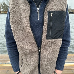 Beige teddyväst med ficka på bröstet - Säljer en mysig beige fleeceväst med dragkedja och en praktisk bröstficka med dragkedja. Perfekt för lager-på-lager under kyliga dagar. Västen har en normal passform och är lätt att matcha med olika outfits.