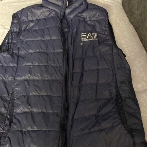 Blå dunväst från Emporio Armani - Snygg blå dunväst från Emporio Armani med EA7-logga på bröstet. Västen har en quiltad design och dragkedja framtill. Perfekt för att hålla värmen med stil. Pris kan diskuteras så hör bara av er 