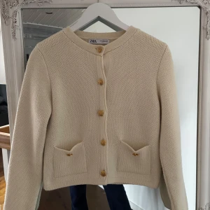 Beige kofta från Zara - Säljer en elegant beige kofta från Zara med guldfärgade knappar och två små fickor framtill. Koftan har en rund halsringning och långa ärmar. Perfekt för en stilren look.