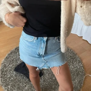 Blå jeanskjol från Zara - Trendig blå jeanskjol från Zara med rå kant och klassisk femficksdesign. Perfekt för en avslappnad stil. Kjolen har en knapp och dragkedja framtill.