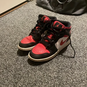 Nike Air Jordans 1 mid storlek 37,5 - Snygga Nike Air Jordans i klassisk färgkombination av svart, röd och vit. Skorna har en hög modell med snörning och den ikoniska Air Jordan-loggan på sidan. Kontakta vid minsta fundering (pris kan diskuteras)