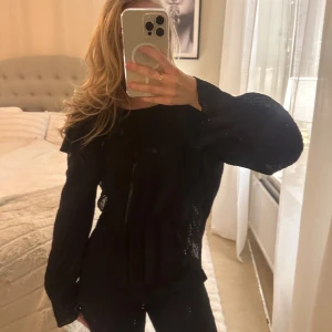 Svart blus med volangdetaljer - Snygg svart blus med volangdetaljer och långa ärmar. Perfekt för en stilren look. Blusen har en elegant design som passar till många tillfällen.