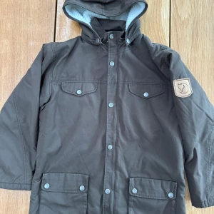 Fjällräven Kids Greenland Winter Jacka Brun - Säljer en brun jacka från Fjällräven med huva och flera fickor med knappar. Jackan har ett mjukt, vitt foder på insidan för extra värme. Perfekt för kalla dagar. Märkeslogga på ärmen ger en snygg touch. 🧥
