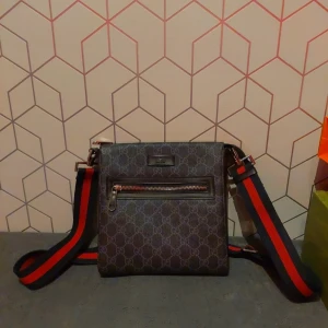 Svart axelväska med mönster - Snygg svart axelväska med ett subtilt mönster och en praktisk dragkedja framtill. Väskan har en justerbar axelrem i blått och rött, vilket ger en cool kontrast. 