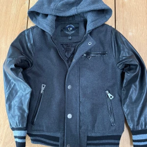 Grå basebolljacka med huva från Urban Republic - Säljer en snygg grå basebolljacka med från Urban Republic med huva och svarta skinnärmar. Jackan har dragkedja och knappar framtill samt flera praktiska fickor med dragkedjor. Perfekt för kyligare dagar med sin quiltade insida.