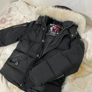  Moose Knuckles - Säljer en svart dunjacka från Moose Knuckles med avtagbar huva och pälsdetalj. Jackan har dragkedja och knappar framtill samt ribbade muddar vid ärmsluten. Perfekt för kalla vinterdagar.