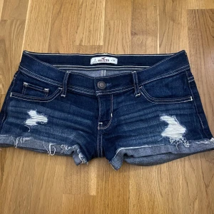 Mörkblå jeansshorts från Hollister - Snygga mörkblå jeansshorts från Hollister med slitna detaljer och fransig kant. Perfekta för en avslappnad stil. De har en knapp och dragkedja framtill samt klassiska fem fickor.
