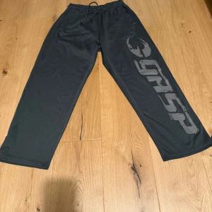 Svarta träningsbyxor från GASP - Säljer ett par svarta träningsbyxor från GASP med elastisk midja och snörning. Byxorna har en lös passform och ett stort tryck på ena benet. Perfekta för gymmet.