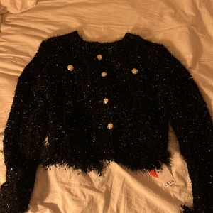 Svart glittrig jacka från Cider - Säljer en svart glittrig jacka från Cider med dekorativa guldknappar. Jackan är croppad och har en unik textur som ger en festlig look. Perfekt för att sticka ut i mängden!
