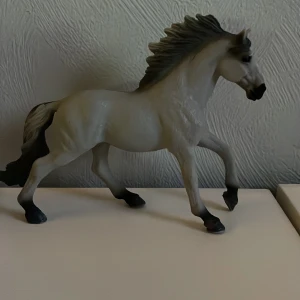 Schleich häst - Detaljerad vit hästfigur i plast med mörk man och svans. Hästen är i en dynamisk pose som ger intryck av rörelse. Perfekt för samlare eller som dekoration.