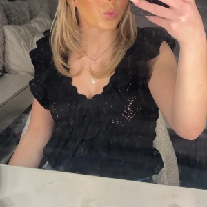 Svart spetsblus - Säljer en elegant svart spetsblus med volangdetaljer. Blusen har en v-ringad design och är ärmlös, vilket gör den perfekt för olika tillfällen. Den är både stilren och bekväm.