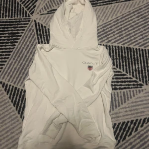 Vit hoodie från GANT - Säljer en stilren vit hoodie från GANT med deras klassiska logga på bröstet. Tröjan har en bekväm passform med långa ärmar och en praktisk magficka. Perfekt för en avslappnad look.(HAR EN BLÅ LITEN FLÄCK) använd 1 gång.