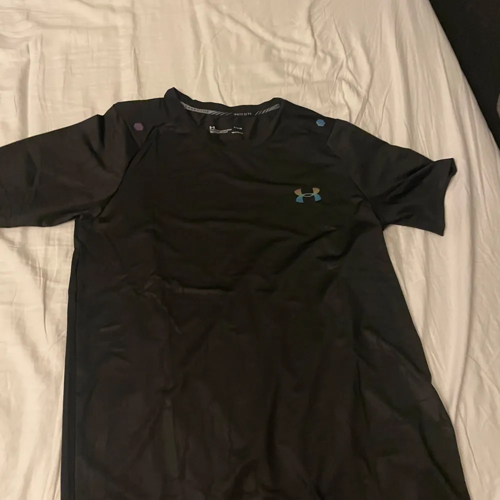 Svart under armour T-shirt med reflekterande logga på bröstet och ett riktigt fett mönster över hela tröjan. Helt oanvänd. Tveka inte att höra av dig vid minsta fundering!. T-paidat.