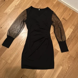 Svart klänning med transparenta ärmar - Snygg svart klänning med transparenta långa ärmar dekorerade med pärlor. Klänningen har en omlott-design framtill och en elegant v-ringning. Perfekt för en stilfull kväll. Aldrig använd.