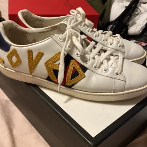 Vita sneakers  - Snygga vita sneakers från Gu**i med texten 'Loved' i guld på sidan. Skorna har röd och blå randdetalj samt röd och blå häl. Perfekta för att ge en lyxig touch till din outfit. Jag får inte skriva märket för plick tar ner annonsen då men de är äkta har bara inget bevis på det