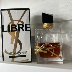 Libre Le Parfum från Yves Saint Laurent - Säljer en elegant flaska av Libre Le Parfum från Yves Saint Laurent. Flaskan har en stilren design med en gyllene YSL-logga och en svart, geometrisk kork. Parfymen har en djup bärnstensfärg som ger en lyxig känsla. Perfekt för den som vill ha en sofistikerad doftupplevelse.
