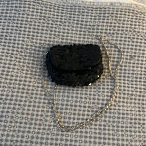 Svart glittrig axelväska med kedja - Snygg svart axelväska med glittriga detaljer och en elegant kedja som axelrem. Perfekt för att ge en extra touch till din outfit. Väskan har en kompakt design och är idealisk för att bära med sig det viktigaste. Väldigt liten men får plats med kort och tuggummi.