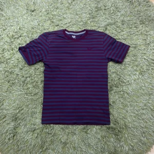 Randig t-shirt från Nike - Säljer en randig t-shirt från Nike i lila och grått. Den har korta ärmar och ett broderat Nike-märke på bröstet. Perfekt för en avslappnad stil.