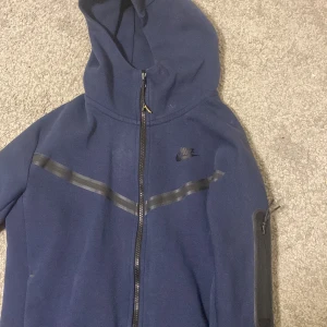 Mörkblå Nike tech fleece Nike - Säljer en mörkblå hoodie från Nike med dragkedja och huva. Jackan har ett diskret Nike-logotyptryck på bröstet och en svart randdetalj över bröstet. Perfekt för en sportig look.finns bara en skada där med Zipen har ändrat färg som ni ser på bilden det guldiga men man kan ta av.Finns några små små hål men man märker ej
