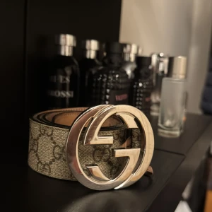 Gucci bälte med GG-logga - Snyggt Gucci bälte med den ikoniska GG-loggan i silver. Bältet har ett klassiskt mönster i beige och brunt, perfekt för att lyfta vilken outfit som helst. Ett måste för modeintresserade!