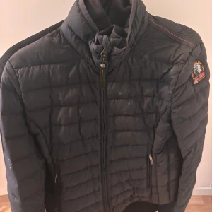 Svart dunjacka från Parajumpers - Säljer en svart dunjacka från Parajumpers med dragkedja och hög krage. Jackan har ett quiltat mönster och en broderad logga på ärmen. Perfekt för kyliga dagar!