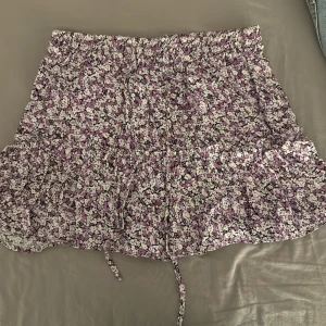 Blommig kortkjol från Zara - Säljer en söt blommig kortkjol från Zara i lila och vit. Kjolen har volanger och ett elastiskt midjeband med knytband för extra passform. Perfekt för en somrig look! 🌸