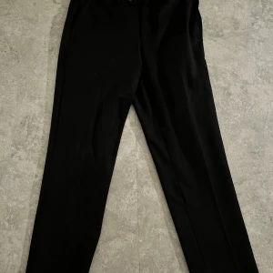 Svarta kostymbyxor från Greve - Eleganta svarta kostymbyxor från Greve. De har en klassisk design med knappar och dragkedja i midjan. Perfekta för en stilren look.