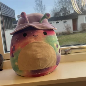 Squishmallow med galaxmönster och hatt - En stor och mjuk Squishmallow med ett färgglatt galaxmönster i lila, rosa, blått och grönt. Den har en söt hatt i lila och antenner som ger den ett unikt utseende. Perfekt för kramar och dekoration.