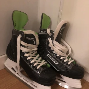 Svarta skridskor från Bauer - Säljer ett par svarta skridskor från Bauer med gröna detaljer. De har snörning och en robust design som passar perfekt för isaktiviteter. Skridskorna har en bekväm passform och är idealiska för både nybörjare och erfarna åkare.
