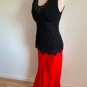 Svart och röd klänning med spetsdetaljer - Elegant klänning med svart överdel och röd kjol. Överdelen har vacker spets och pärldetaljer, medan kjolen är lång och flödande. Perfekt för en speciell kväll.