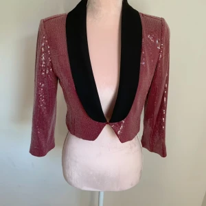 Paljettprydd croppad kavaj - Snygg och glittrig kavaj med paljetter i både rosa och grått. Den har en djup v-ringning och en elegant svart krage. Kavajen är croppad och har trekvartsärmar, perfekt för en festlig look.