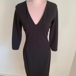 Svart klänning med v-ringning - Elegant svart klänning med v-ringad hals och långa ärmar. Perfekt för en stilren look. Klänningen har en figurnära passform som framhäver silhuetten.