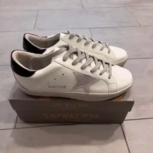 Säljer ett par snygga Golden Goose sneakers i vitt med grå stjärndetalj och svart häl. Skorna har en cool vintage-look med snörning och är i mycket bra skick. Perfekta för både vardag och fest! Box och Kvitto ingår.