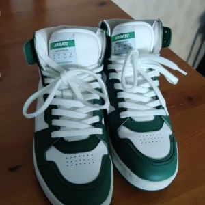 Grönvita sneakers från Axel Arigato St39 - Säljer ett par snygga sneakers från Axel Arigato i grönt och vitt. De är i mycket bra skick och har en cool hög modell med snörning. Perfekta för både vardag och fest! Tillverkade i Portugal med högkvalitativt material. Storleken på skorna är 39 eu. Priset kan diskuteras