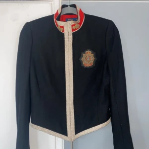 Svart kavaj från Polo Ralph Lauren - Säljer en elegant svart kavaj från Polo Ralph Lauren med guldiga detaljer och ett broderat emblem på bröstet. Kavajen har en unik design med röd krage och guldfärgade knappar. Perfekt för höst och vinter, och ger en stilren look till både vardag och fest. Anser att den är i strl XS trots att beskrivning anger S. 