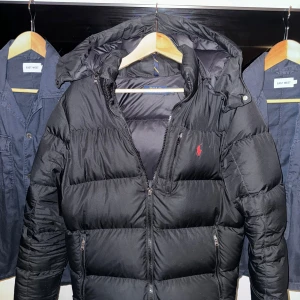 Ralph Lauren dunjacka - Säljer en snygg svart pufferjacka från Ralph Lauren. Jackan har en dragkedja framtill och en mysig huva. Perfekt för kalla dagar med sin vadderade design. Tyvärr har en bit från ena dragkedjan gått av men den funkar fortfarande som den ska (inget man tänker på om man inte tittar noga) men lågt pris på grund av defekten. Kan verifiera äktheten med kvitto från NK, hör av er om ni undrar något :) Pris kan diskuteras 