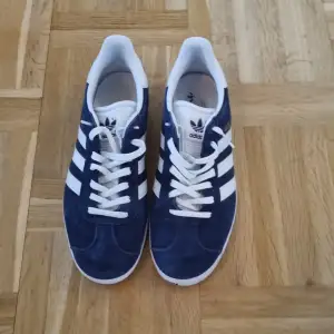 Säljer ett par snygga blå Adidas Gazelle sneakers i mocka. De har de klassiska vita ränderna och snörning. Perfekta för både vardag och fest! Skorna är i mycket bra skick och redo för en ny ägare.