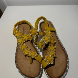Gula sandaler med blommor och pärlor - Snygga gula sandaler med blommiga detaljer och pärlor. Perfekta för sommaren med en bekväm passform och elastisk rem. Sandalerna har en glittrig kant som ger extra stilpoäng. Perfekta för både vardag och fest!