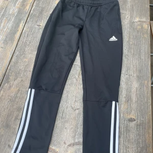 Svarta träningsbyxor från adidas - Säljer ett par svarta träningsbyxor från adidas. De har en elastisk midja och vita ränder längs benen. Perfekta för träning eller en avslappnad dag. Byxorna är i bra skick och säljes för endast 20 kr. 