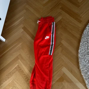 Röda mjukisbyxor från Nike - Säljer ett par snygga röda mjukisbyxor från Nike. De har en cool randig detalj längs sidorna och en vit Nike-logga på låret. Perfekta för en avslappnad stil eller träning. Byxorna har en bekväm passform med resår i midjan och snörning för justering.