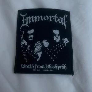 Svart tygmärke med Immortal-motiv - Säljer ett svart tygmärke med motiv från bandet Immortal. Texten 'Wrath from Blashyrkh' är broderad i vitt tillsammans med bandets logga och ett coolt mönster. Perfekt för att pimpa din jacka eller väska med lite metal-stil! 🤘