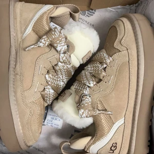 Beige lowmel uggs - Säljer ett par snygga och bekväma sneakers från UGG i beige mocka. De har en fluffig vit insida som håller fötterna varma och breda snören i ett coolt mönster. Perfekta för höst och vinter, med en robust sula för bra grepp. Passar både till vardags och lite mer avslappnade tillfällen. (Säljer utan förpackningen!)