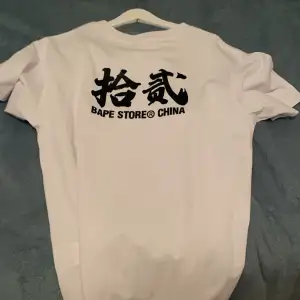 Säljer en vit t-shirt från Bape Store China med ett coolt blått och vitt mönster på framsidan. T-shirten har korta ärmar och är i bomull, perfekt för en avslappnad stil. Text och grafik på baksidan ger den en unik touch. Passar perfekt till vardags eller en chill dag ute.