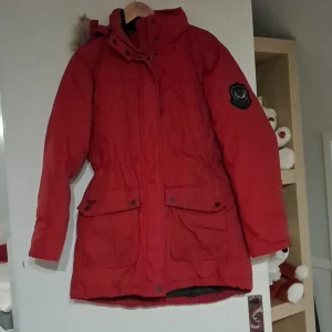 Röd parkajacka med pälsdetalj - Säljer en snygg röd parkajacka med pälsdetalj på huvan. Jackan har långa ärmar och praktiska fickor framtill. Perfekt för kalla vinterdagar och höstpromenader. Den har en skön passform och är i bra skick. Perfekt för dig som vill hålla stilen i kylan!