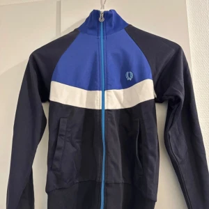 Blå och svart jacka från Fred Perry - Säljer en snygg blå och svart jacka från Fred Perry i storlek 34. Jackan har en dragkedja framtill och är tillverkad av 50% bomull och 50% polyester. Perfekt för en sportig look med sitt klassiska märke på bröstet. Passar bra till både vardag och träning!